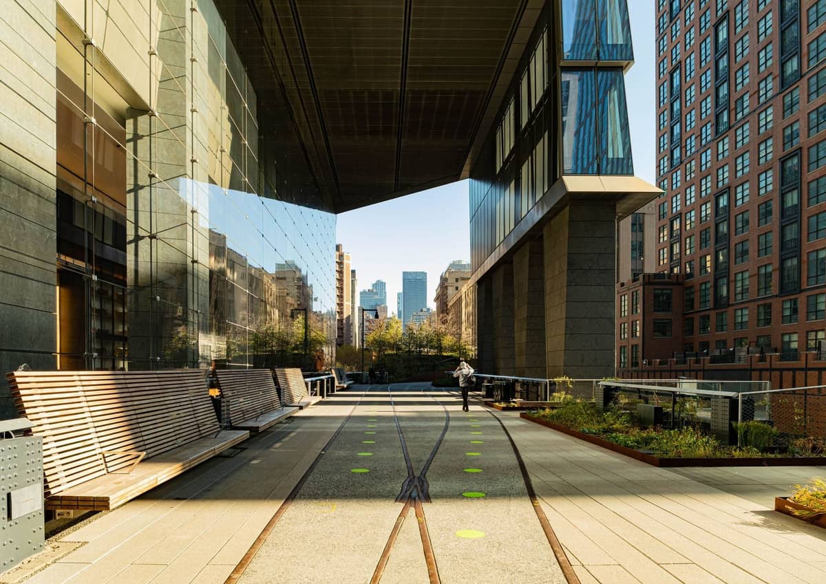 Blick entlang des High Line Parks in Manhattan mit Bänken, Grünflächen und modernen Gebäuden, die den erhöhten Gehweg unter sonnigem Himmel einrahmen.