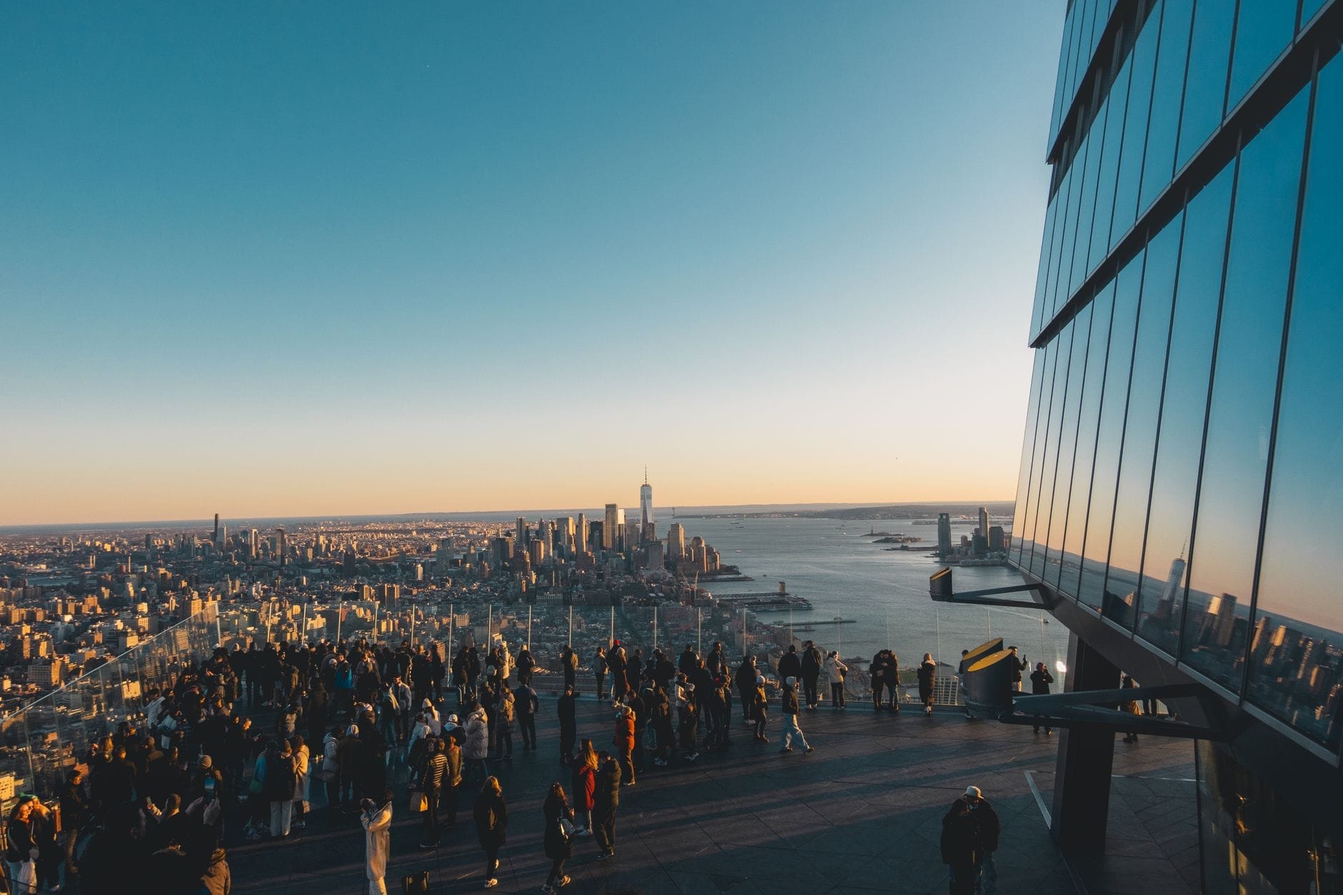 Grande multidão aproveitando a vista panorâmica de Manhattan e do One World Trade Center em um moderno mirante externo ao pôr do sol, com o skyline da cidade e o Rio Hudson ao fundo.