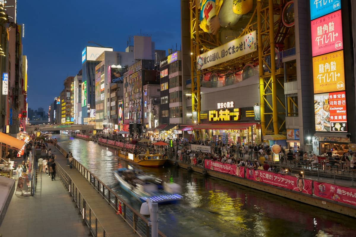 Evening view of Dotonbori canal in Osaka with neon-lit buildings, busy waterfront walkways, and boats gliding on the water.