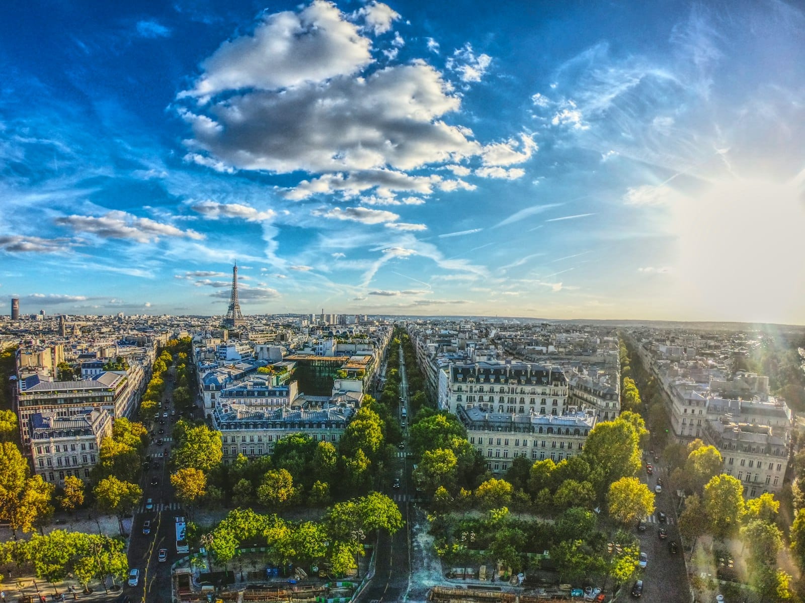 Una spettacolare vista a volo d'uccello di Parigi sotto un cielo azzurro intenso con nuvole sparse e la Torre Eiffel visibile in lontananza, con la luce del sole che diffonde un bagliore dorato sui viali e sui tetti della città.