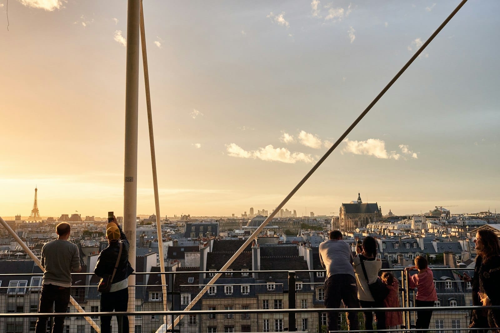 Menschen auf einer Dachterrasse in Paris bei Sonnenuntergang, die über die Dächer der Stadt blicken, mit dem Eiffelturm in der Ferne.