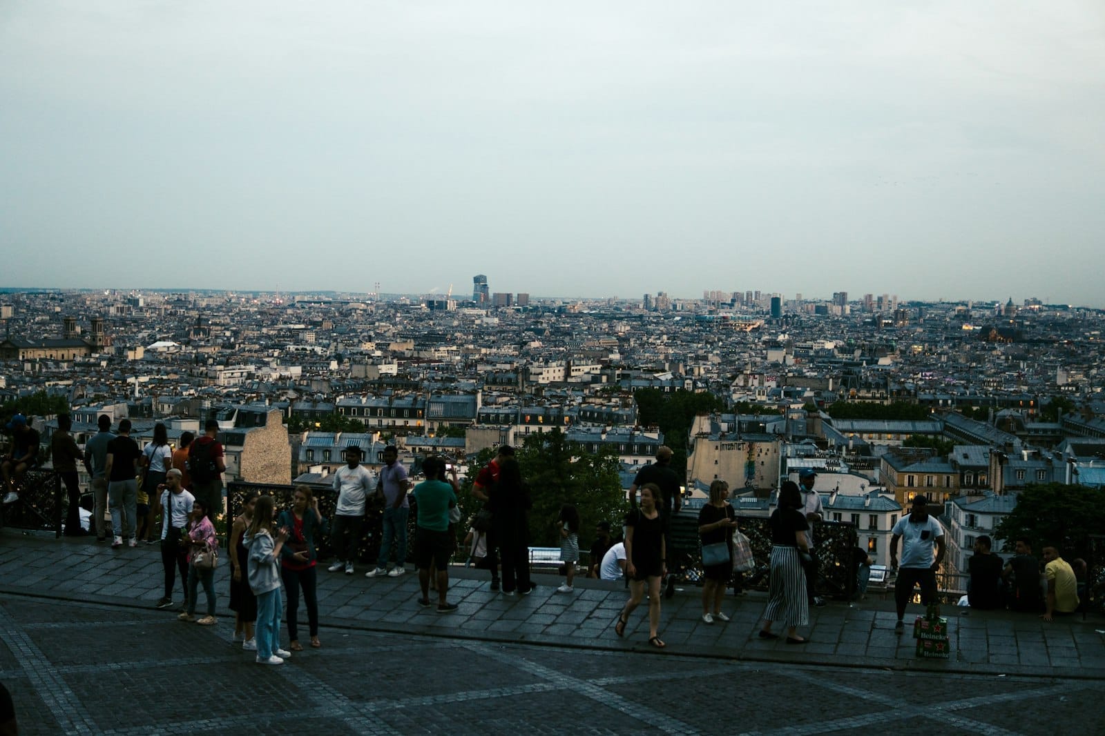Blick vom Sacré-Cœur mit Menschengruppen auf der Esplanade, die bei Dämmerung die Stadtsilhouette von Paris überblicken.