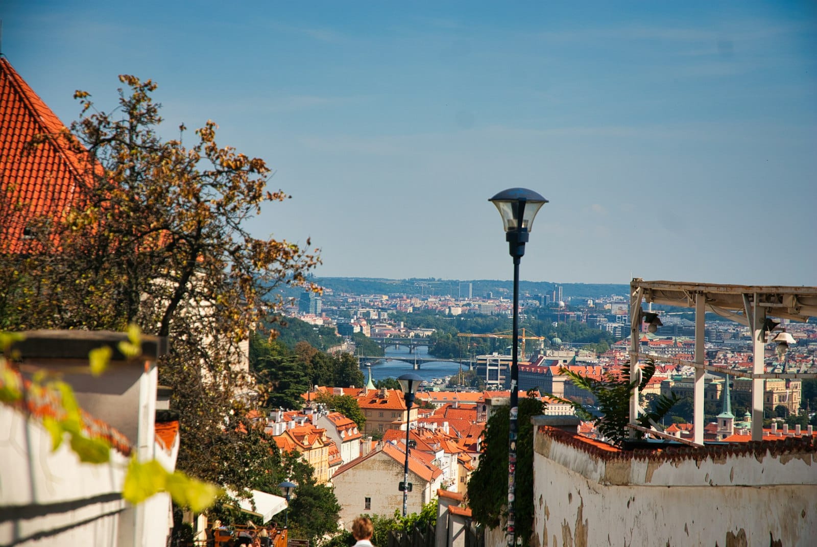 Prag Kalesi bölgesinin içindeki bir terasydan kırmızı çatılara, uzakta Vltava Nehri'ne ve mavi gökyüzü altında şehir siluetine bakan manzara.