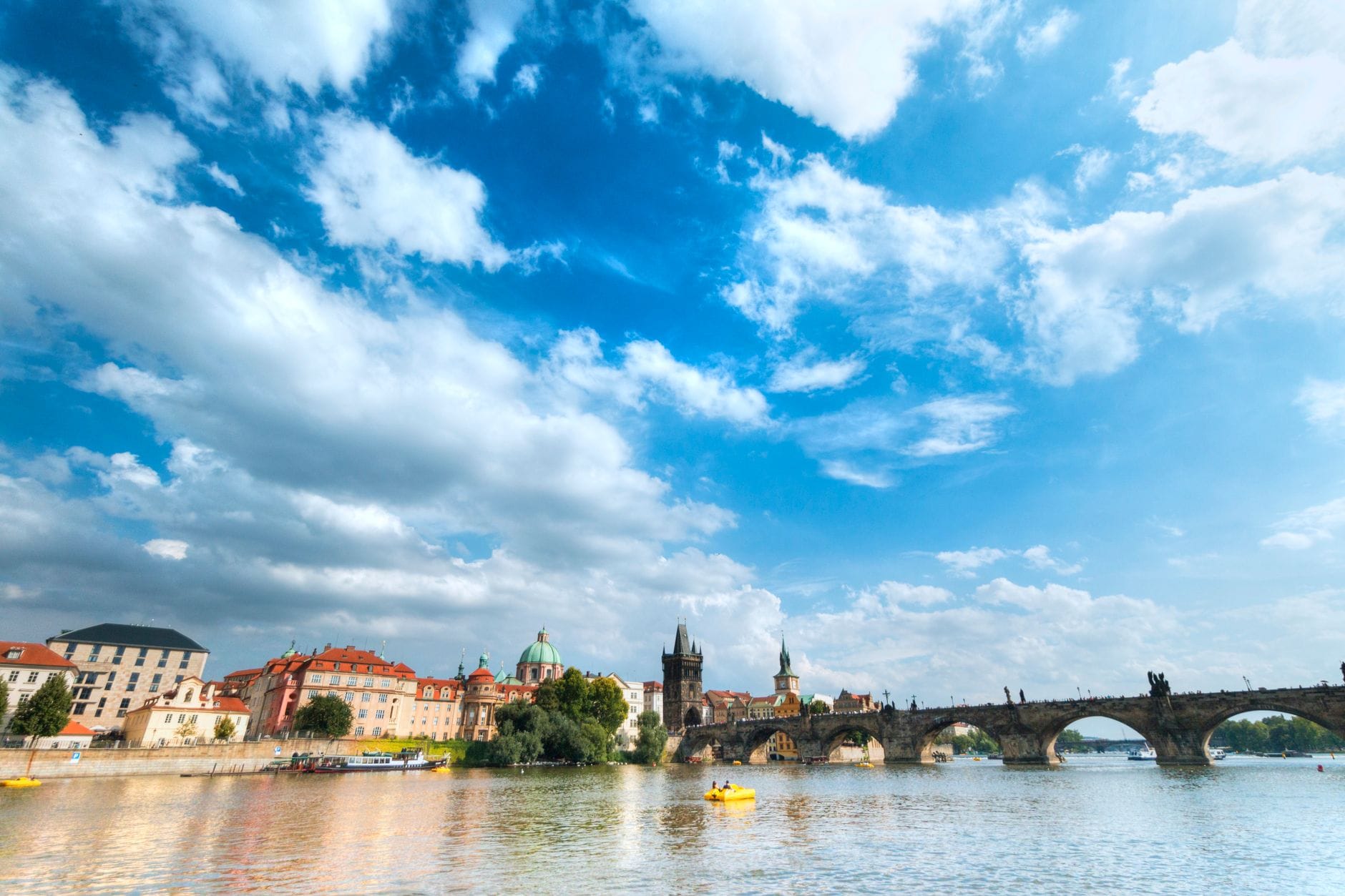 Vltava Nehri'nde kaykalarla su seviyesinden Charles Köprüsü'nün geniş görünümü, tarihi Prag silüeti ve bulutlarla dramatik mavi gökyüzü.