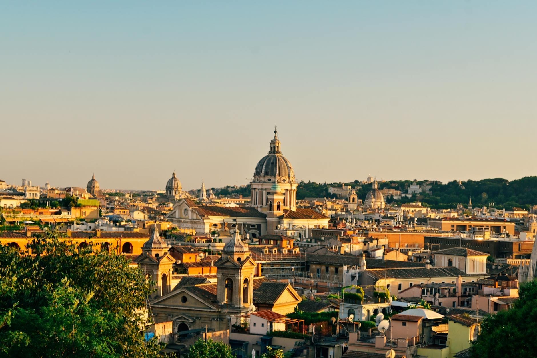 Wide panoramic view of Rome’s rooftops and domes at sunset, seen from a high vantage point with greenery in the foreground.