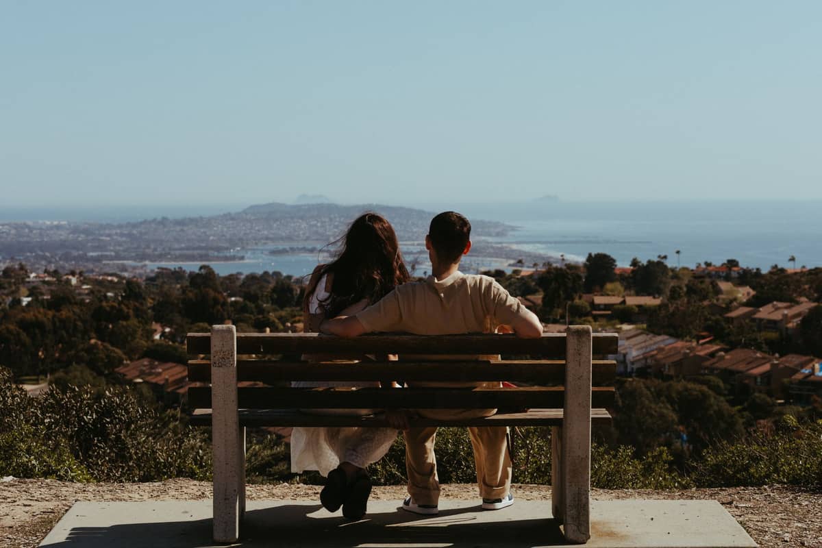 คู่รักนั่งบนม้านั่งบนยอดเขาที่มองเห็นวิวพาโนรามาของชายฝั่งและเมือง San Diego ภายใต้ท้องฟ้าแจ่มใส