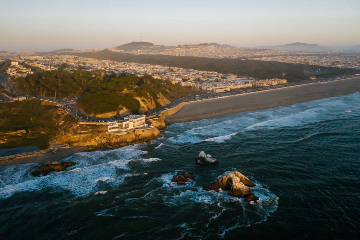 Luftaufnahme von Ocean Beach und Cliff House in San Francisco, wo die zerklüftete Küstenlinie auf den Pazifischen Ozean trifft, mit der Stadtsilhouette und dem Sutro Tower im Hintergrund.