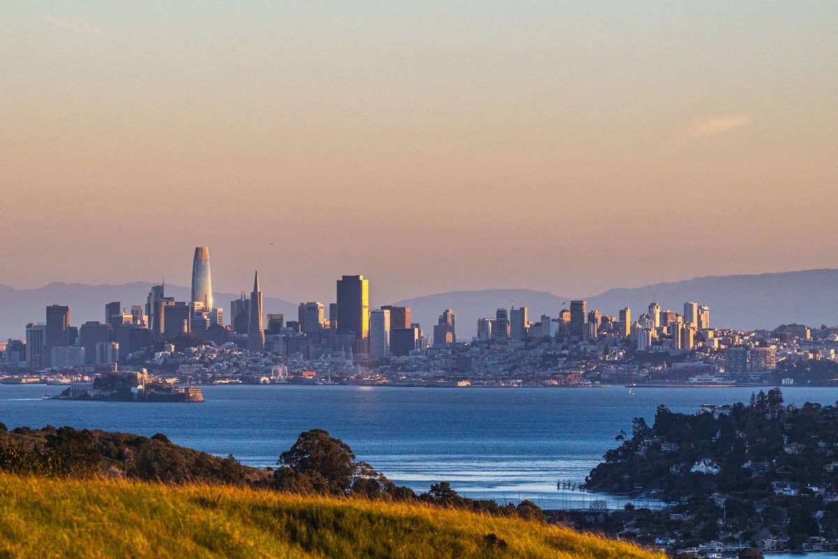Panoramablick auf die Skyline von San Francisco mit den Wolkenkratzern der Innenstadt, der Bucht, Alcatraz Island und sanft geschwungenen grünen Hügeln im Vordergrund bei Sonnenuntergang.