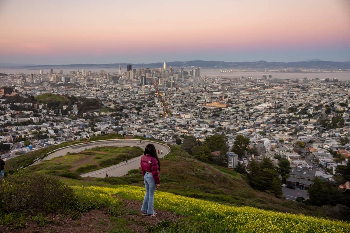 Person auf dem Gipfel der Twin Peaks mit Blick auf die Skyline von San Francisco, kurvenreicher Straße und Panoramablick auf die Stadt bei Sonnenuntergang