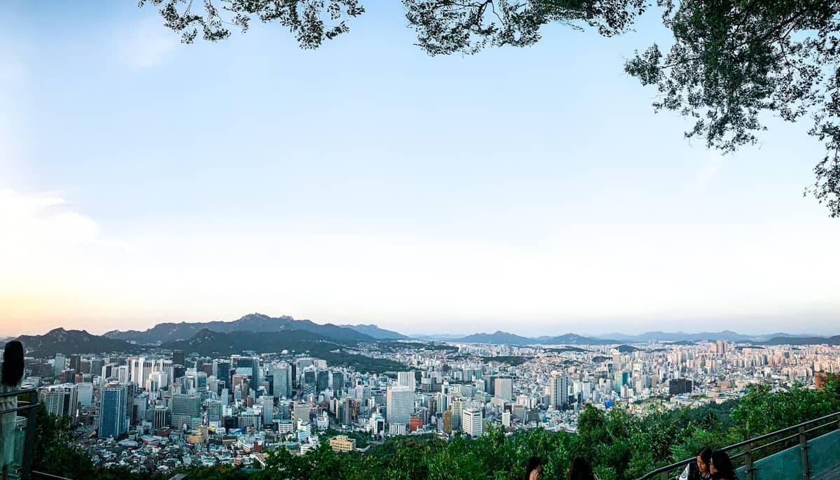Vista panorâmica do centro de Seul a partir de um parque no topo de uma colina, com o skyline da cidade emoldurado por árvores e pessoas aproveitando o mirante.