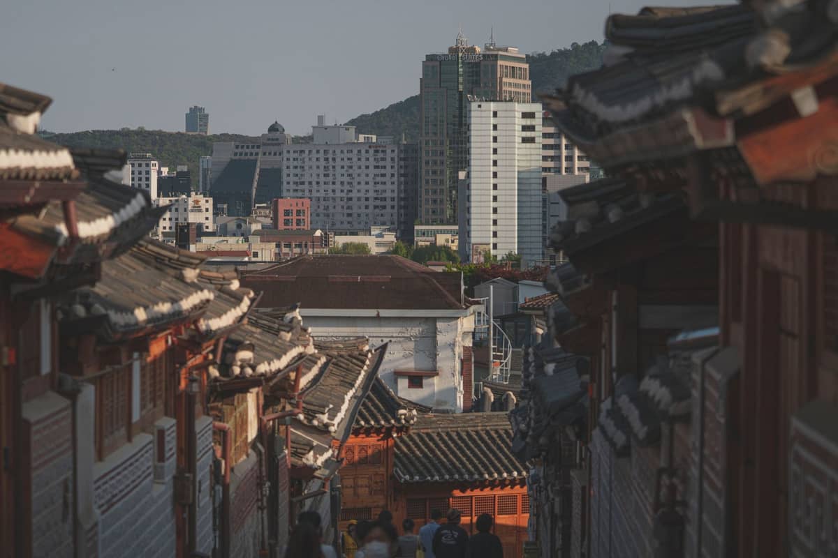 Uma viela inclinada em Seul ladeada por telhados tradicionais hanok, edifícios modernos e colinas distantes à luz do dia.