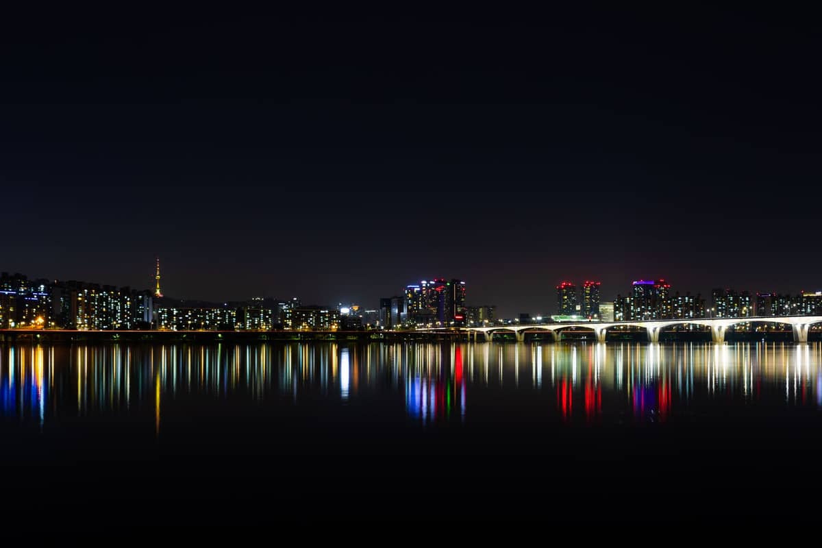 Vista ampla do horizonte noturno de Seul com arranha-céus, reflexos coloridos no rio Han e uma ponte bem iluminada.