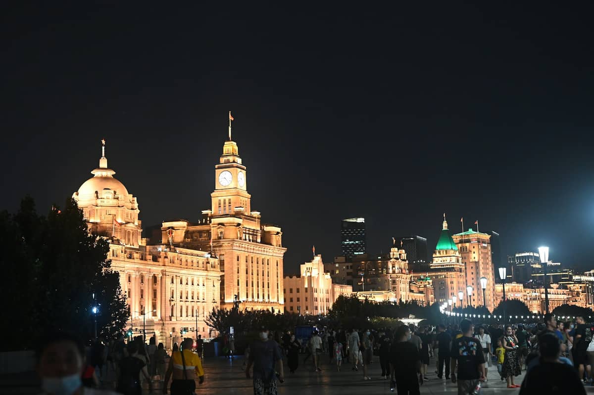 Pemandangan malam promenade Bund di Shanghai dengan gedung-gedung bersejarah yang terang dan orang-orang berjalan di tepi sungai.