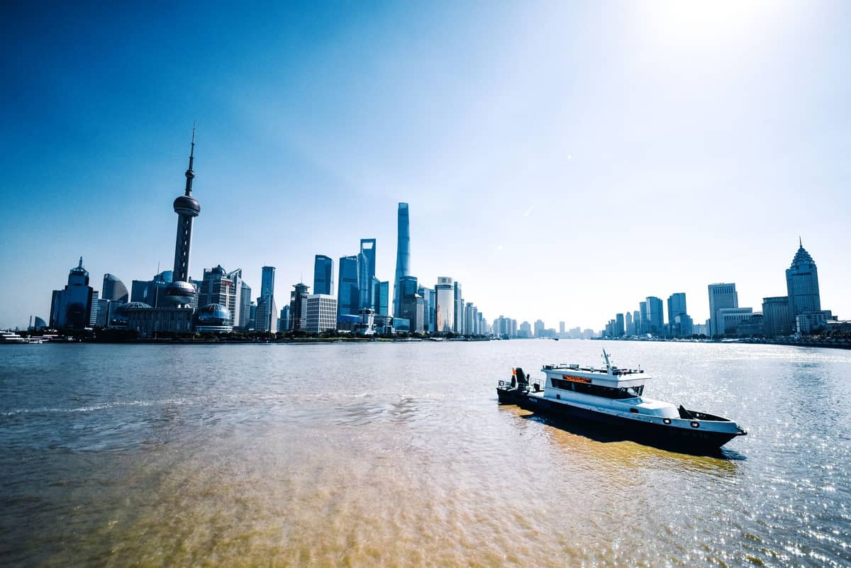 Pemandangan luas melintasi sungai menuju cakrawala Shanghai, dengan sebuah perahu di latar depan dan gedung pencakar langit ikonik yang terlihat di kejauhan.