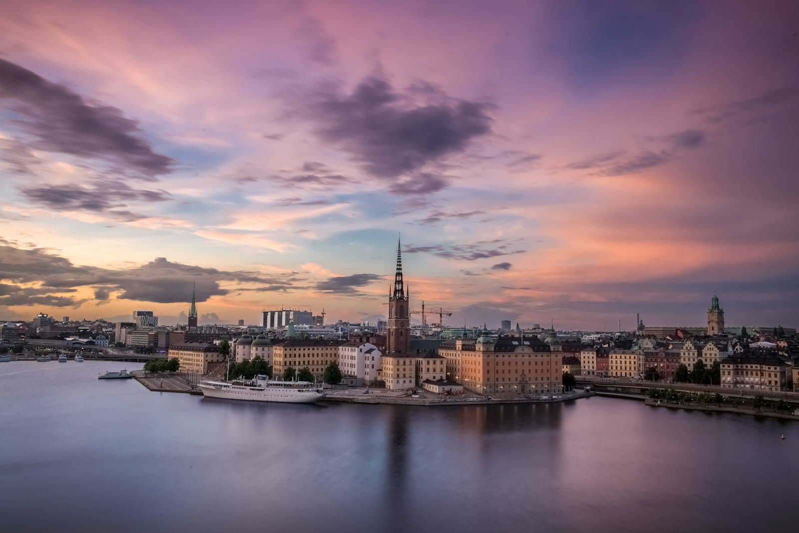 วิวเมืองสตอกโฮล์มยามเย็นจากมุมสูง เห็นโบสถ์ Riddarholmen และอาคารประวัติศาสตร์สีสันสดใคร่ริมแม่น้ำ ใต้ท้องฟ้าสุดดรามาติก