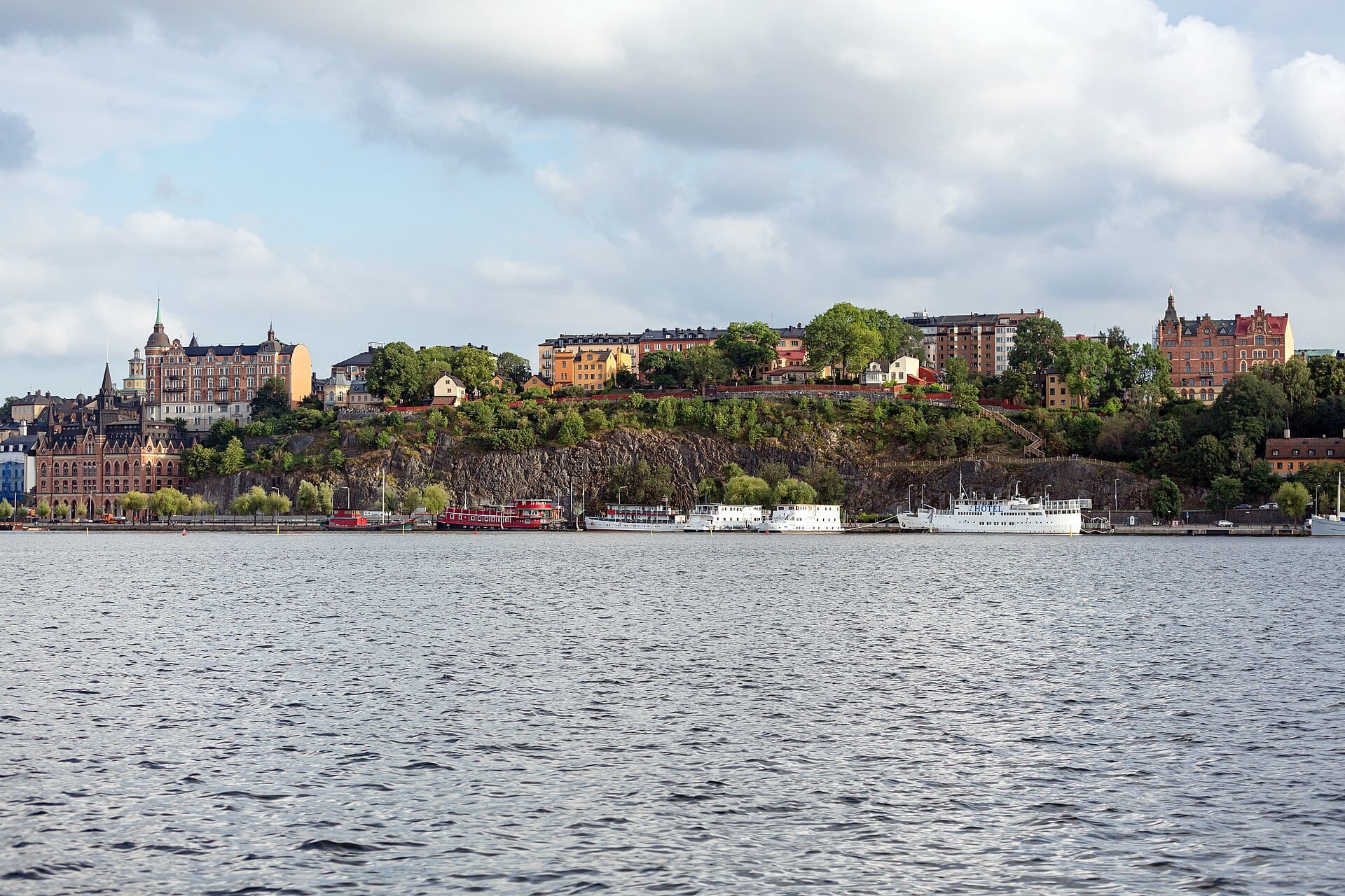 วิวกว้างของขอบหน้าผา Södermalm ในสตอกโฮล์ม พร้อมอาคารสีสันสดใสบนยอด ต้นไม้เขียวชอุ่ม และเรือเรียงรายตามแนวน้ำ