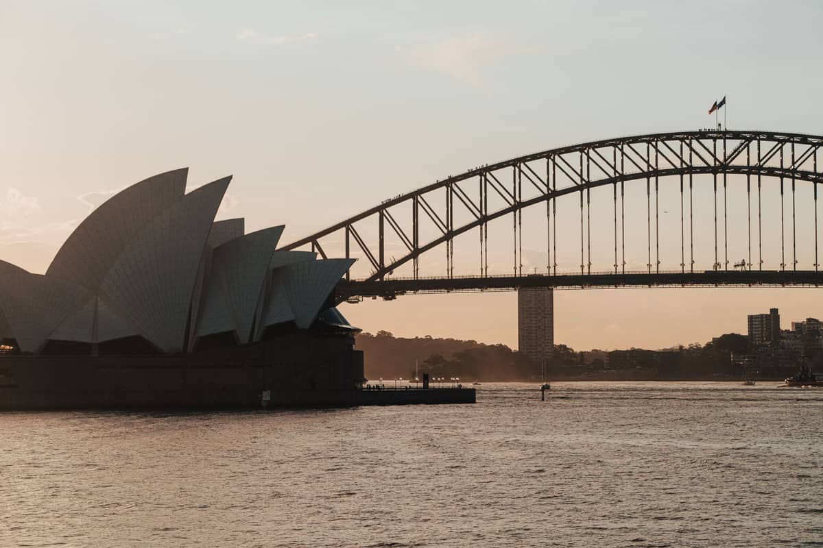Sydney Opera House and Harbour Bridge viewed at sunset across the harbor, with soft golden sky and water in the foreground.