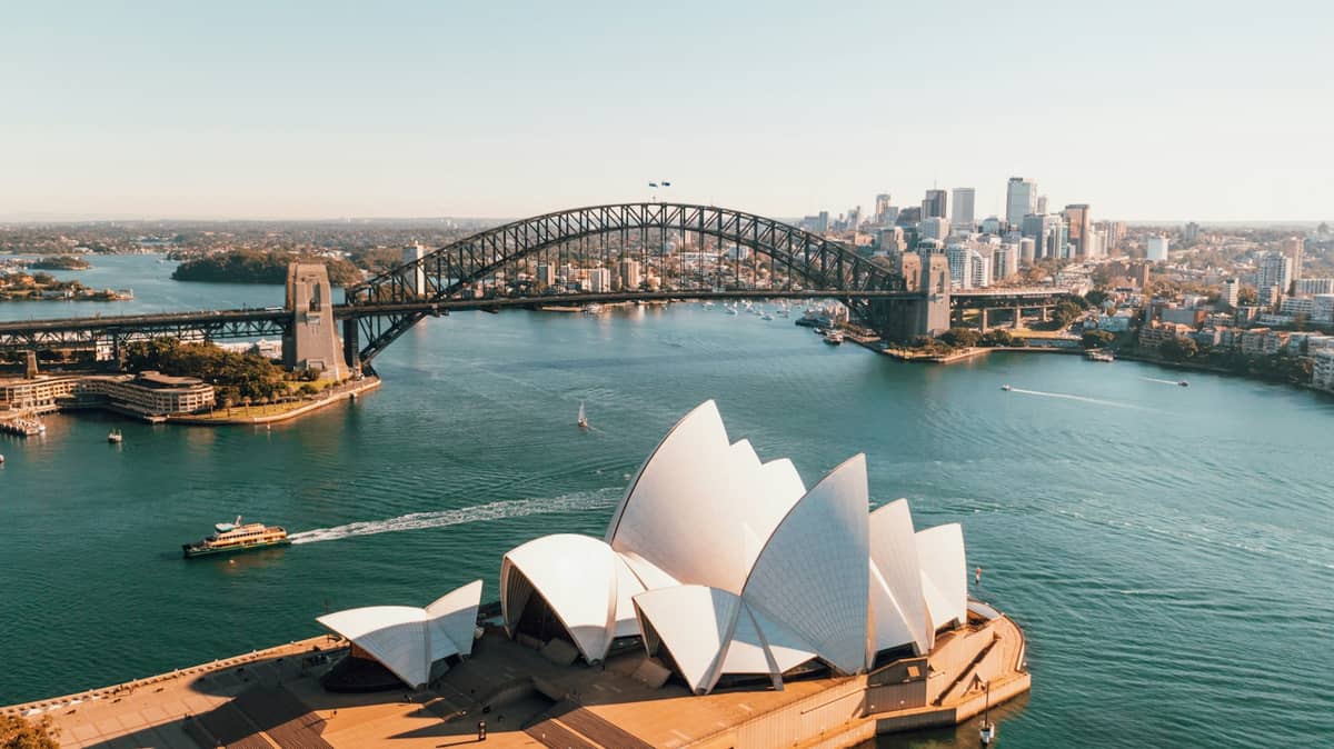 Szeroka panorama z lotu ptaka na zatokę Sydney Harbour z Operą na pierwszym planie, mostem Harbour Bridge i sylwetką miasta w tle, w świetle dnia.