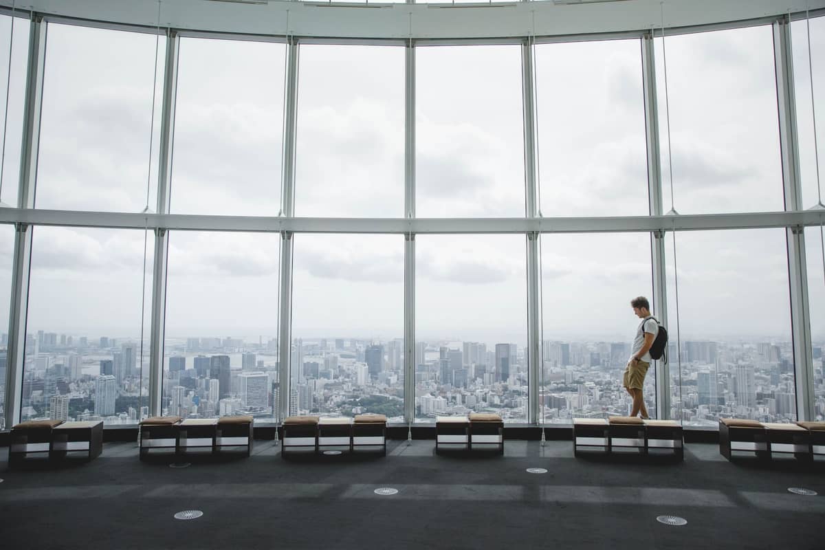 Spacious, modern observation deck with panoramic floor-to-ceiling windows overlooking the Tokyo cityscape, a single visitor enjoying the sweeping daytime urban view.