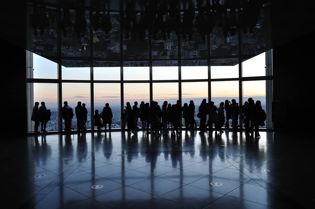 Visitors silhouetted against panoramic windows at a high-rise Tokyo observation deck, gazing out over the city skyline at sunset with reflections on the polished floor.