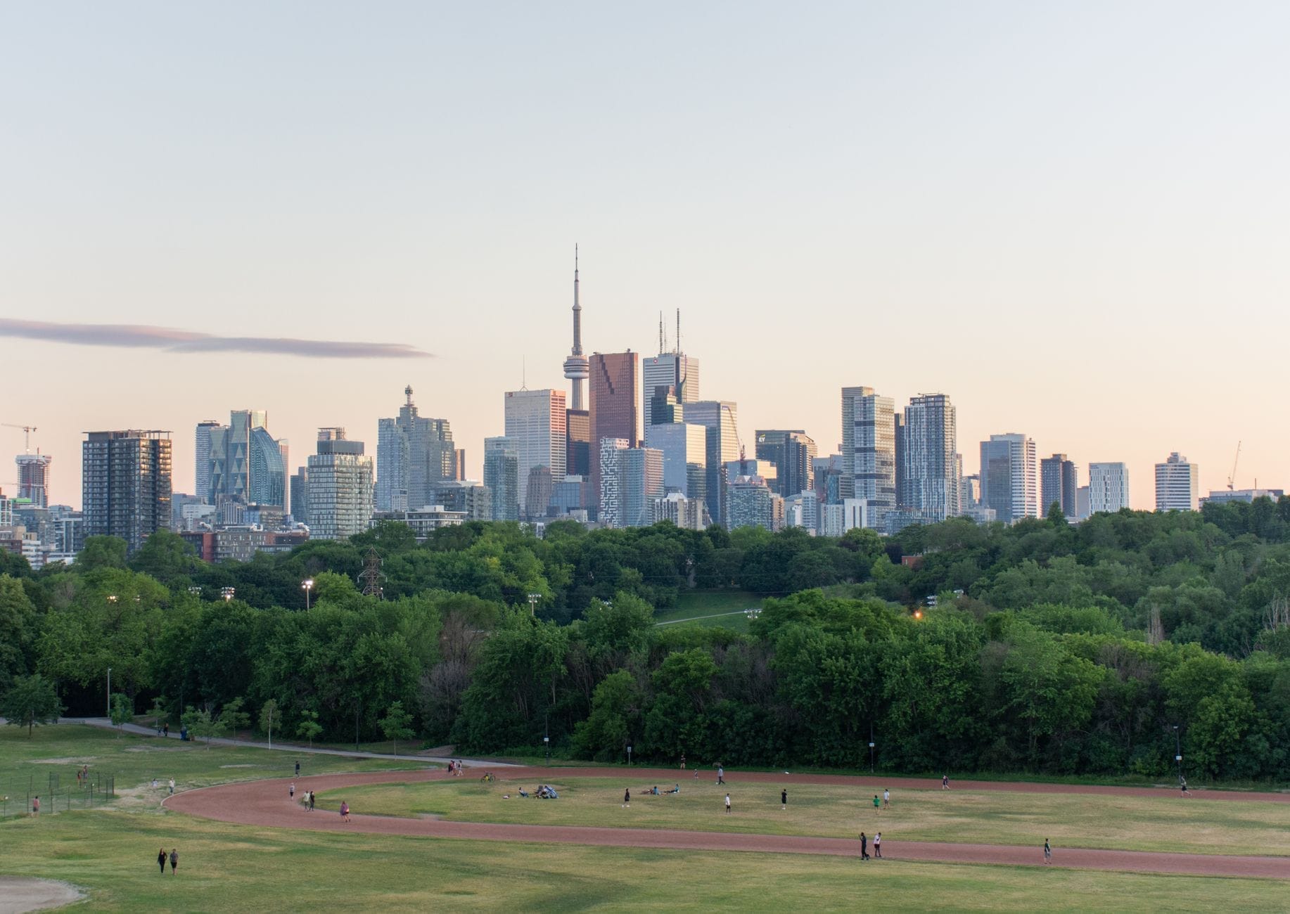 Vista ampla a partir de um parque gramado no topo de uma colina olhando sobre as árvores em direção ao horizonte de Toronto, com a CN Tower e os edifícios da cidade visíveis ao entardecer.