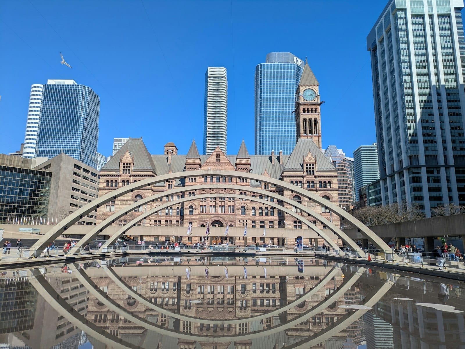 A Nathan Phillips Square de Toronto com seus icônicos arcos, espelho d'água, torre do relógio do Old City Hall e modernos arranha-céus sob um céu azul claro.