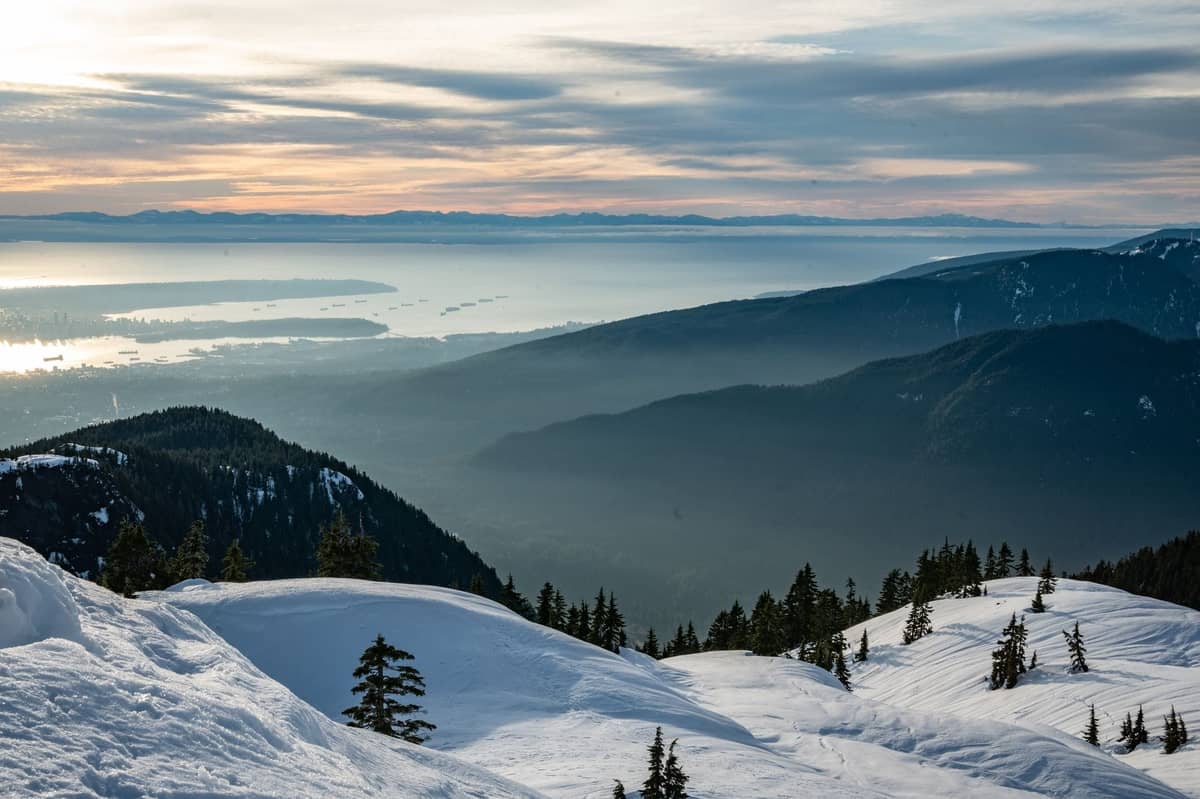 Verschneiter Berggipfel-Aussichtspunkt in den North Shore Mountains mit weitem Panoramablick über Vancouver, das Wasser und die ferne Skyline der Stadt bei Sonnenuntergang.