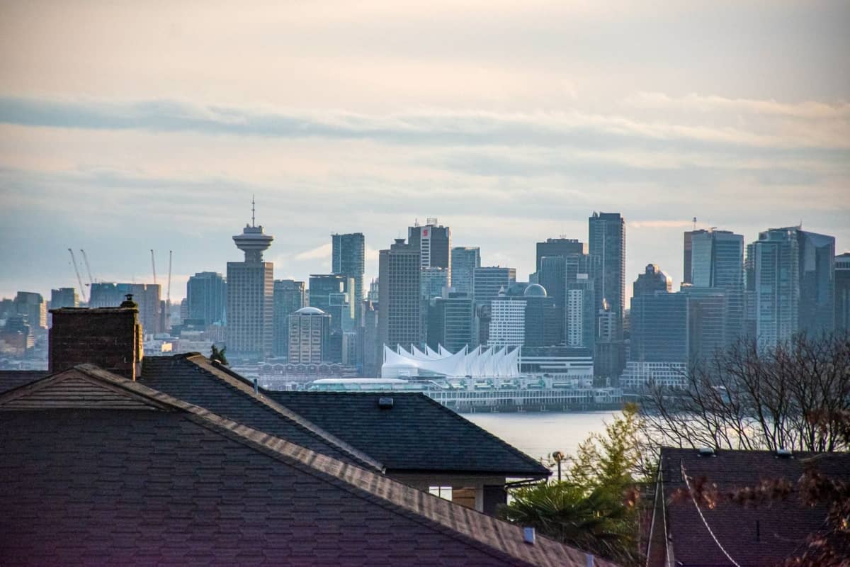 Blick auf die Skyline von Vancouver von einem erhöhten Aussichtspunkt in einem Vorort, mit den Hochhäusern der Innenstadt und Canada Place, die bei Sonnenauf- oder -untergang über den Dächern sichtbar sind.