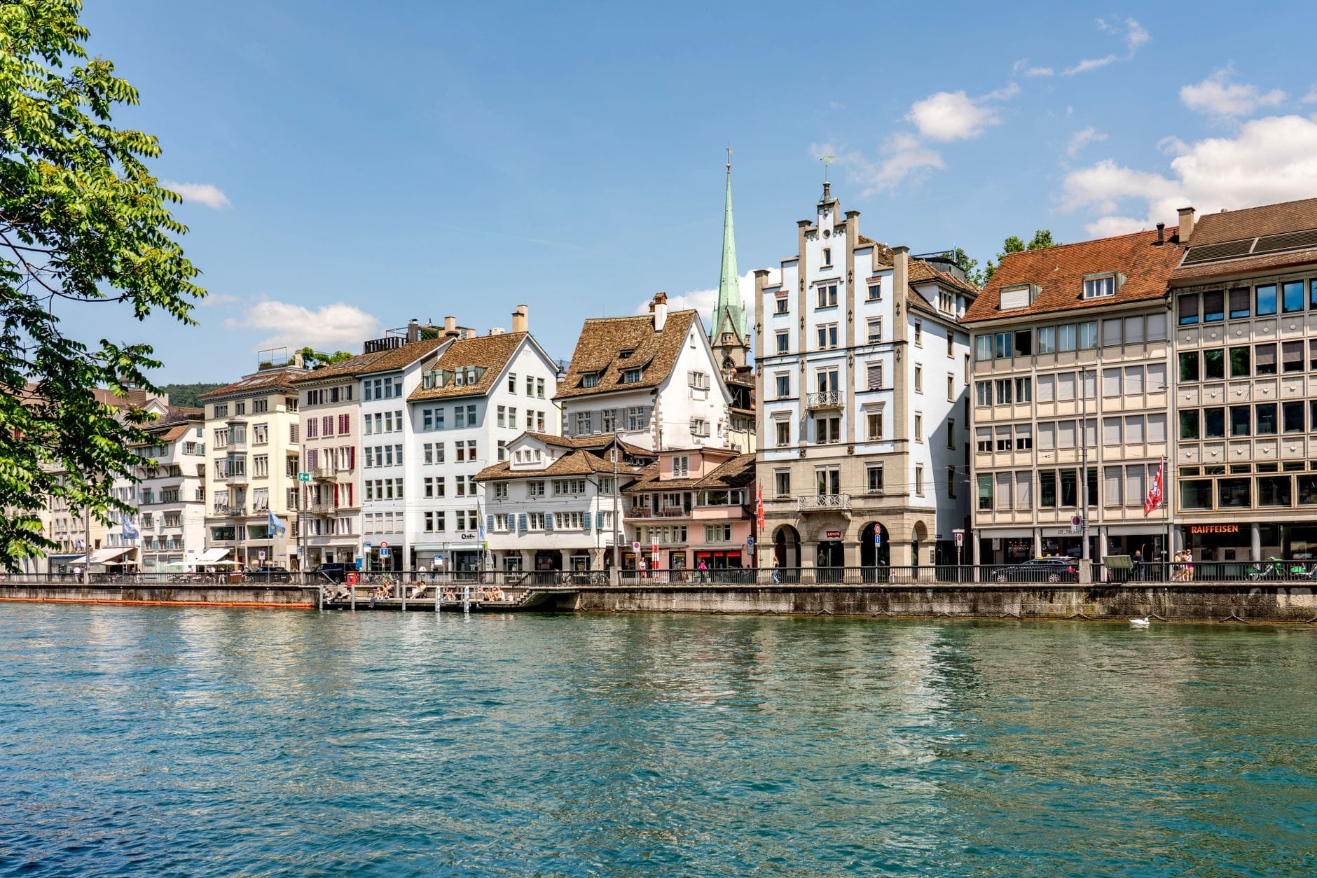 อาคารประวัติศาสตร์ที่มีด้านหน้าสไตล์ยุคกลางริมแม่น้ำ Limmat ในเมือง Zurich โดยมียอดแหลมของโบสถ์ Fraumünster สูงตระหง่านเหนือสถาปัตยกรรมระดับถนน