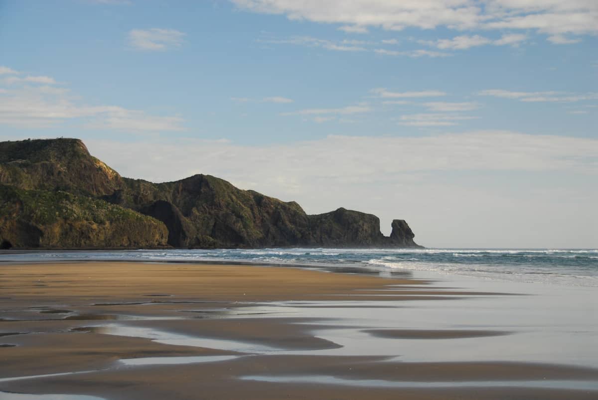 Weitblick auf den Bethells Beach mit schwarzem Sand, Gezeitentümpeln und schroffen grünen Klippen unter einem teils bewölkten blauen Himmel.