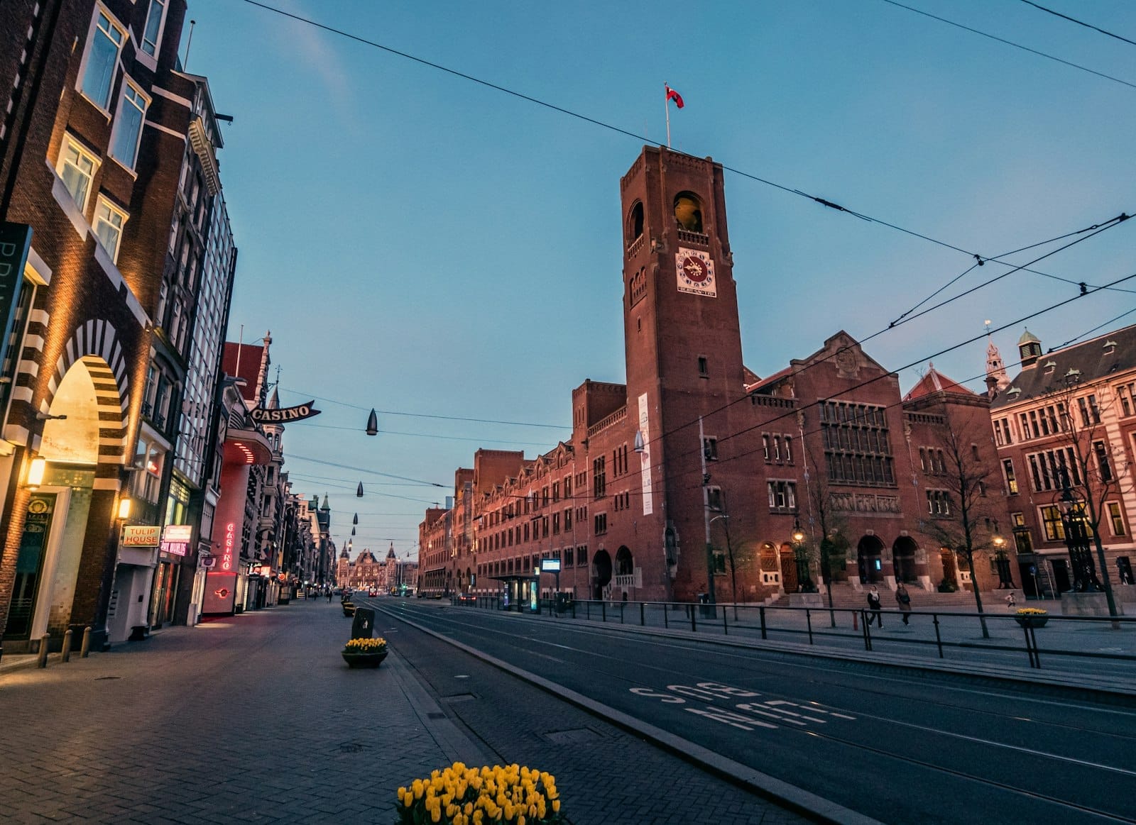 Vista gran angular de la fachada de ladrillo y la torre del reloj del Beurs van Berlage en la calle Damrak, con raíles de tranvía y tulipanes amarillos en primer plano al atardecer.