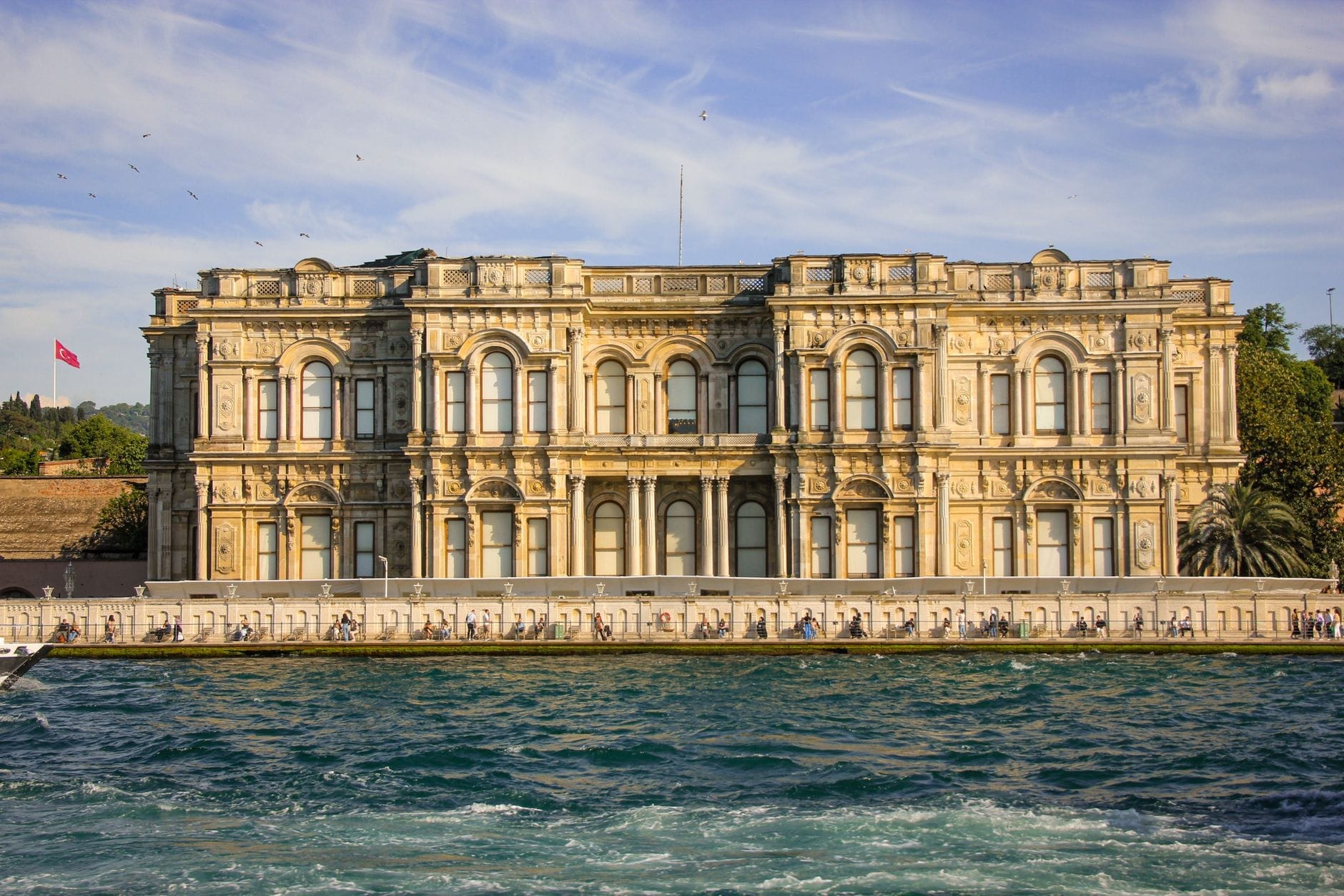Il Palazzo di Beylerbeyi visto dall'altra sponda del Bosforo, con la sua ornata facciata neoclassica, l'acqua azzurra in primo piano e una lussureggiante vegetazione sullo sfondo sotto un cielo luminoso.