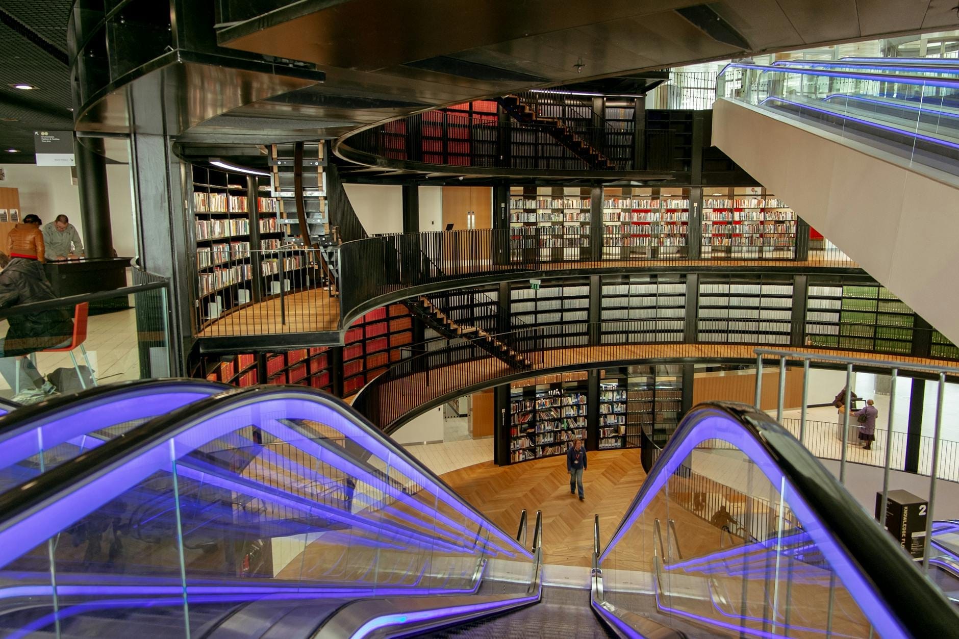 Vista interna moderna di una biblioteca su più livelli con scale mobili ampie, ringhiere in vetro e scaffali che rivestono le pareti circolari.