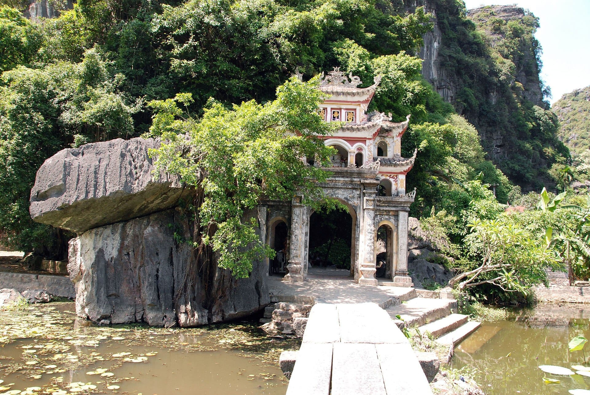 Bích Động Pagoda entrance surrounded by lush greenery, limestone cliffs, and a lotus-filled pond under bright daylight.