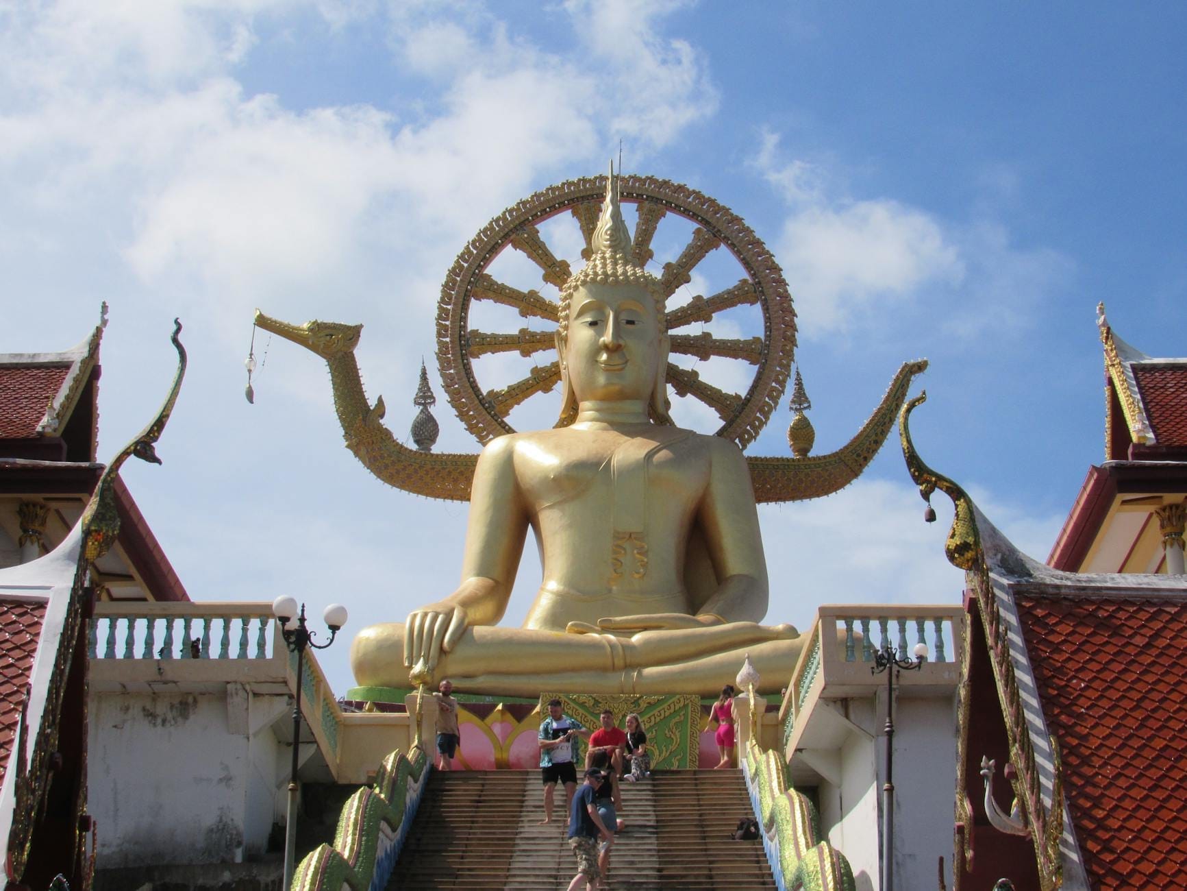 Grande statua del Buddha dorato in cima a una scalinata aperta sotto un cielo azzurro, con i tetti del tempio e i visitatori che salgono i gradini.