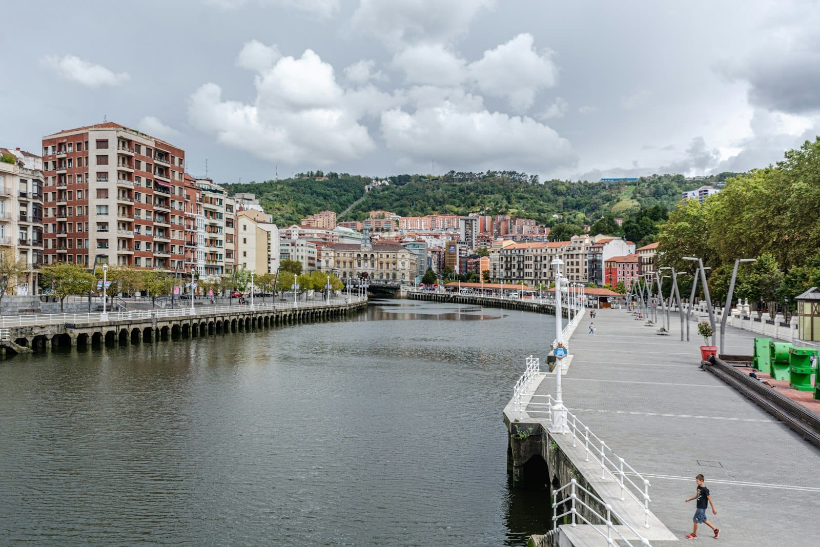 Uferpromenade in Bilbao mit modernen Apartmentgebäuden, Hügeln im Hintergrund und Menschen, die auf dem breiten Gehweg spazieren.