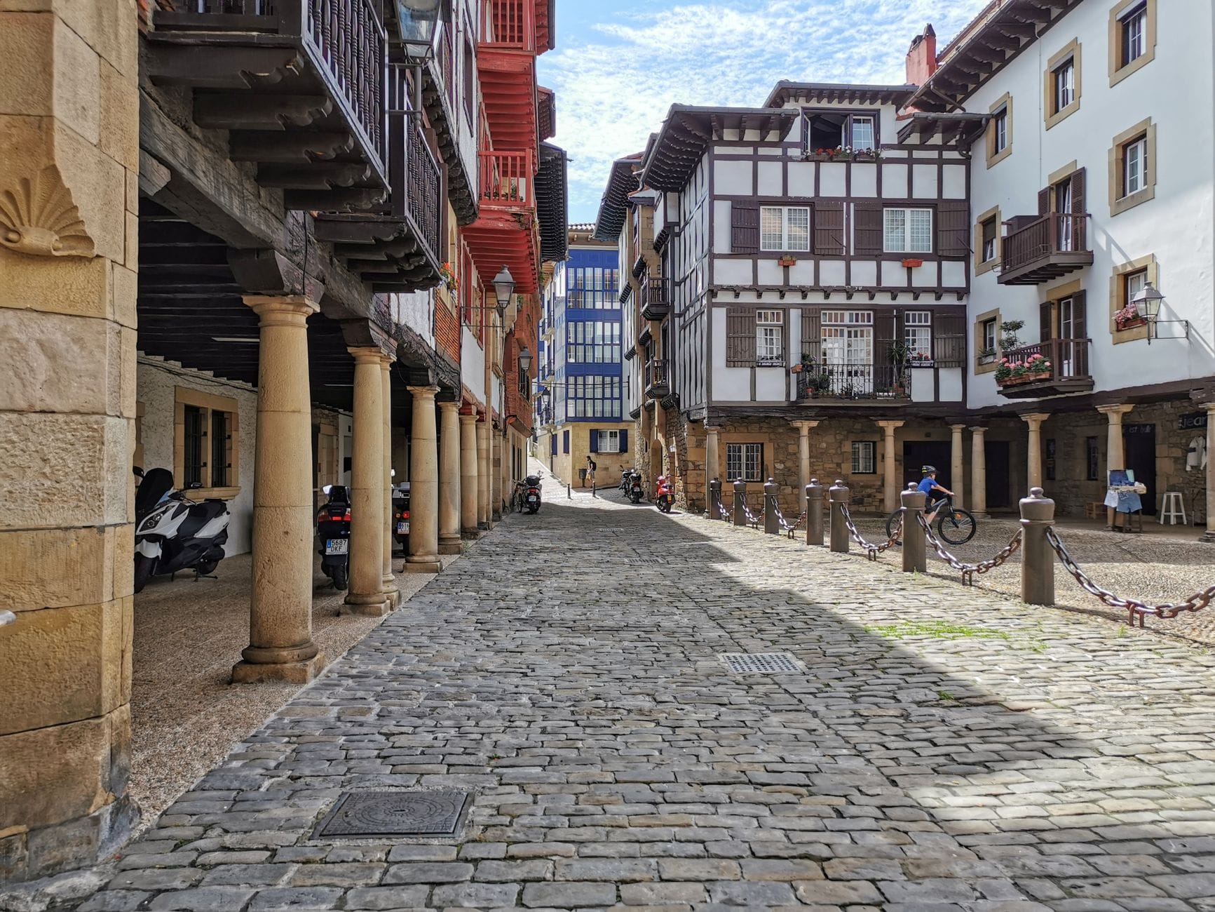 Kopfsteinpflasterstraße gesäumt von historischen Fachwerkhäusern, Steinsäulen und Holzbalkonen in der Altstadt von Bilbao an einem klaren Tag.