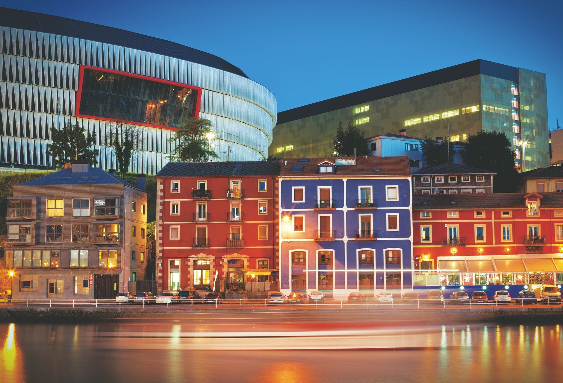 Abendliche Stadtansicht von Bilbao mit bunten Gebäuden am Flussufer, moderner Architektur und einem Stadion im Hintergrund.