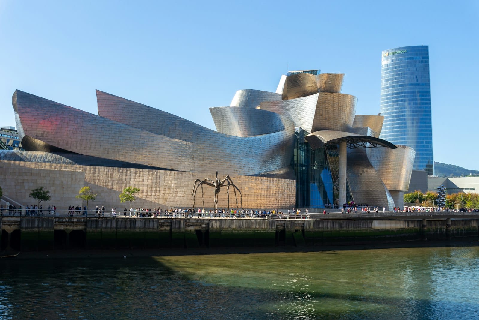 Das Guggenheim Museum Bilbao mit seinen geschwungenen Titanpaneelen, der Lage am Fluss, der Spinnenskulptur und klarem blauem Himmel bei Tageslicht.