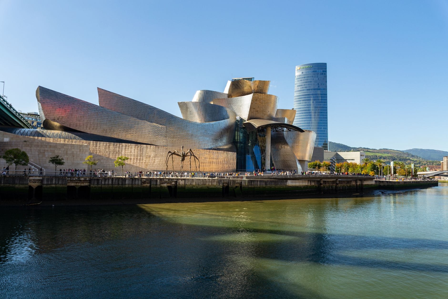 Tagesansicht des Guggenheim Museum Bilbao, Flussufer, moderner Wolkenkratzer und ikonische Spinnen-Skulptur unter einem klaren blauen Himmel.