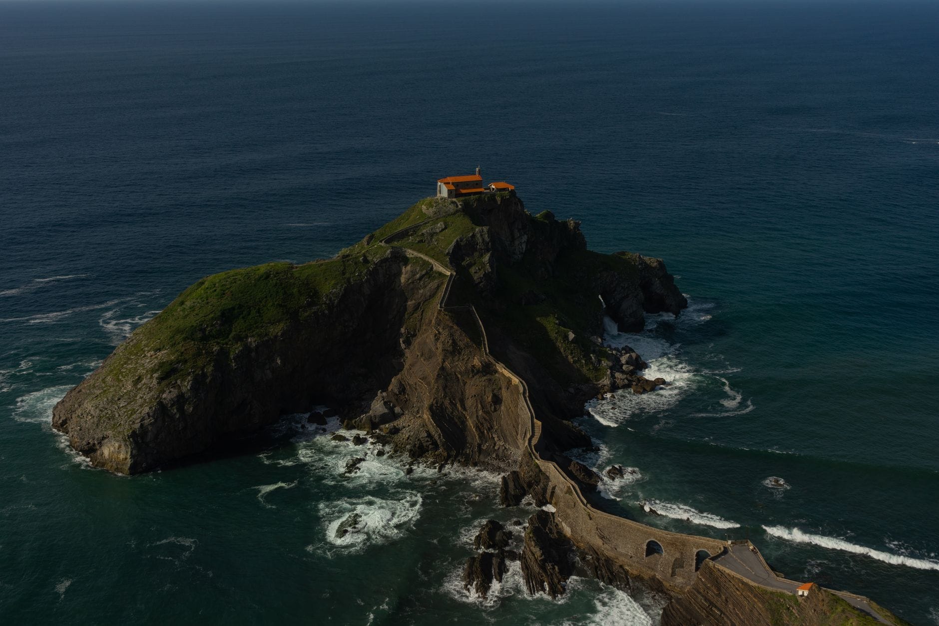 Eine dramatische Luftaufnahme des Felseneilands San Juan de Gaztelugatxe und seiner Steinbrücke, umgeben vom Ozean, mit Wellen, die an der felsigen Küste brechen.