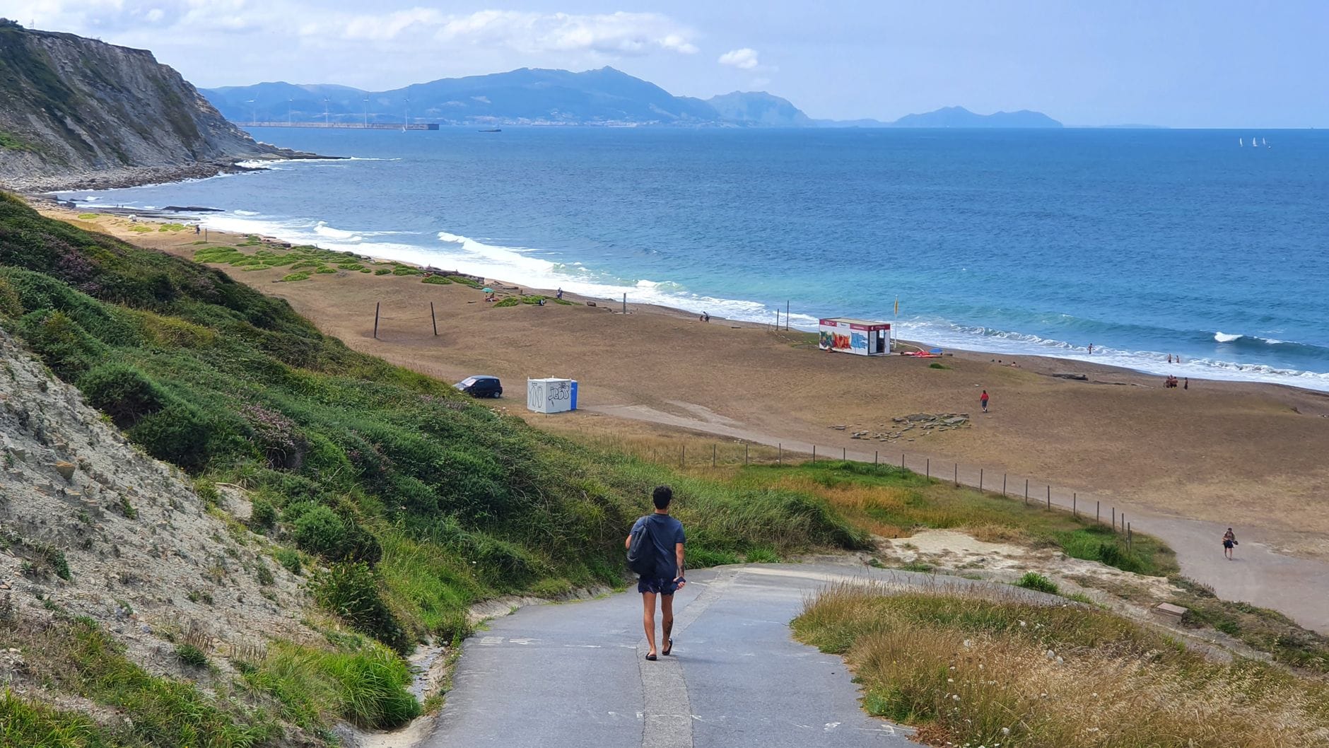 Person, die einen Weg entlang zu einem Sandstrand geht, mit dem Meer und grünen Hügeln im Hintergrund in der Nähe von Bilbao.