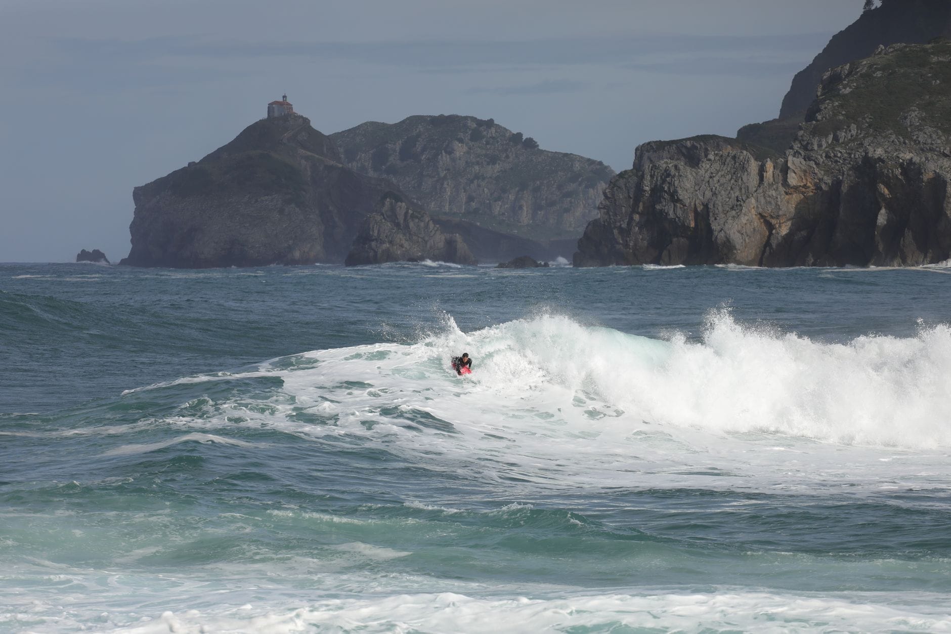 Surfer auf einer Welle nahe dramatischer Klippen mit einer kleinen Kapelle auf der Kuppe im Hintergrund an der Küste von Urdaibai.