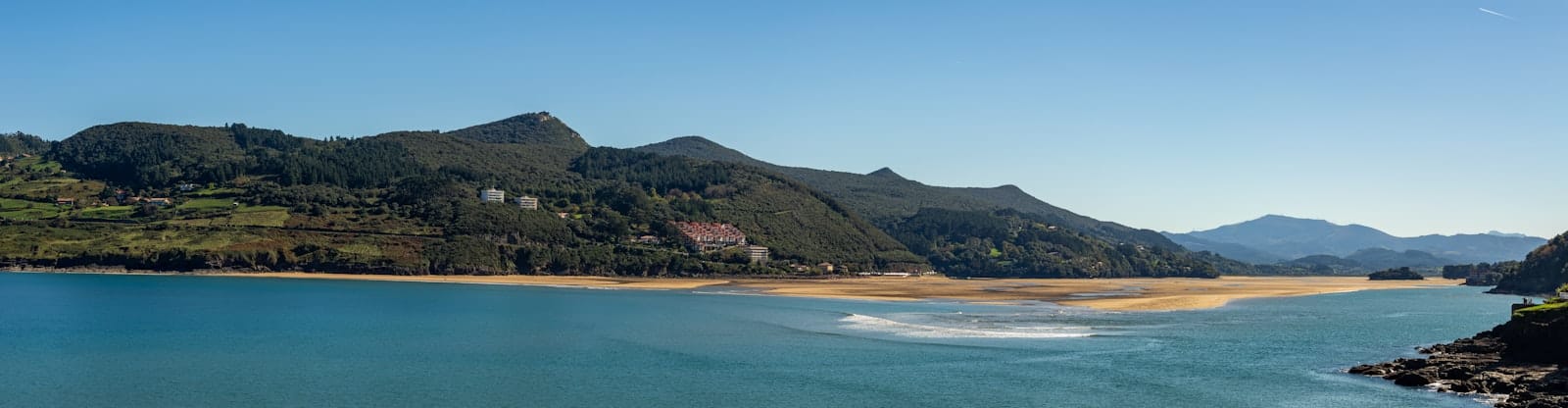 Weitläufiger Panoramablick auf einen Sandstrand mit türkisblauem Wasser, bewaldeten Hügeln und klarem Himmel an der Baskenküste bei Bilbao.
