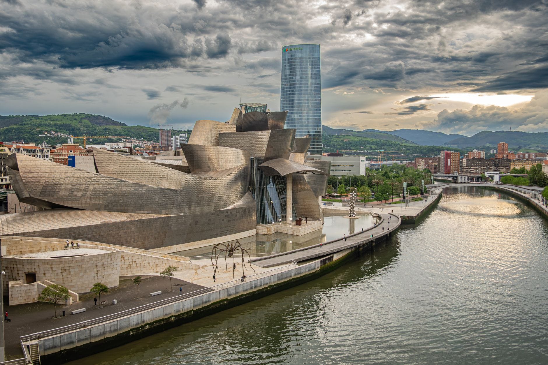 Bilbao’s iconic Guggenheim Museum with its modern architecture beside the river, city skyline and distant mountains under a dramatic cloudy sky at sunset.