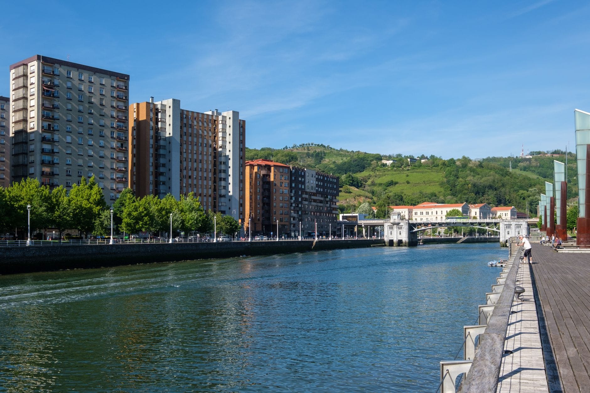 Blick auf die Flusspromenade von Bilbao, moderne Gebäude und grüne Hügel unter einem klaren Sommerhimmel mit wenigen Besuchern.