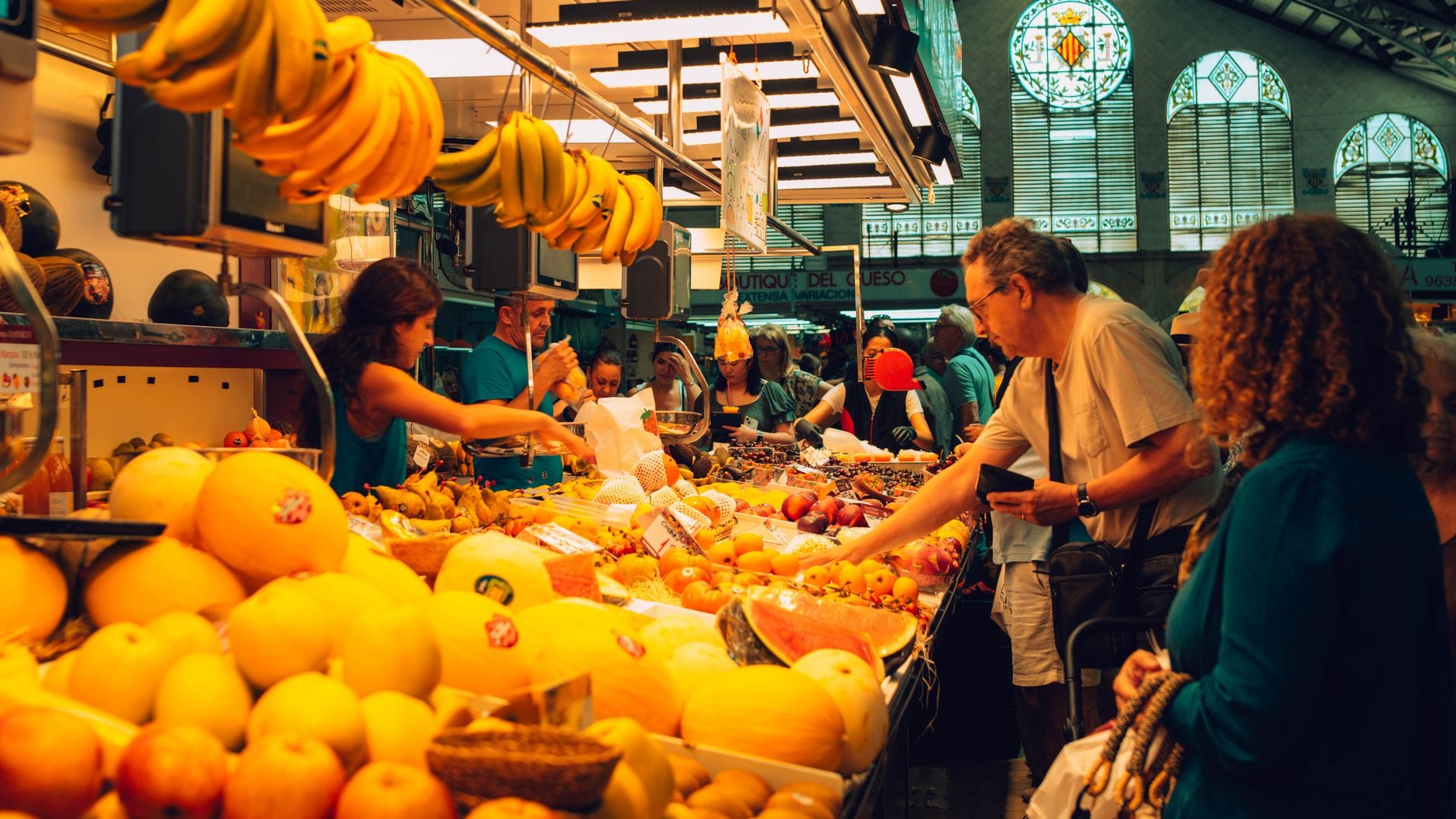 Einkäufer kaufen frisches Obst auf einem lebhaften überdachten Markt in Bilbao mit bunten Buntglasfenstern und hängenden Bananenbündeln über ihnen.