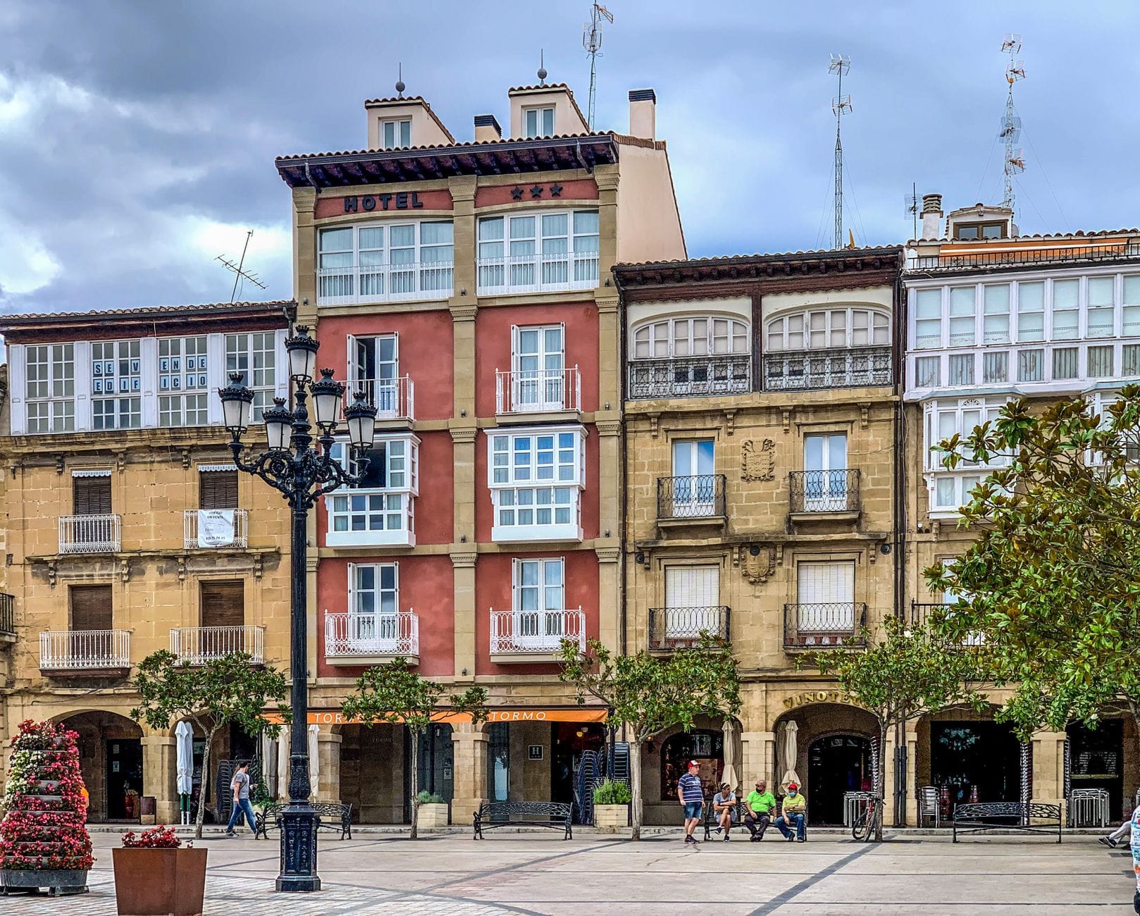 Traditioneller Platz in Bilbao mit einem kleinen Hotel, historischen Fassaden, Bäumen und vorbeilaufenden Menschen.
