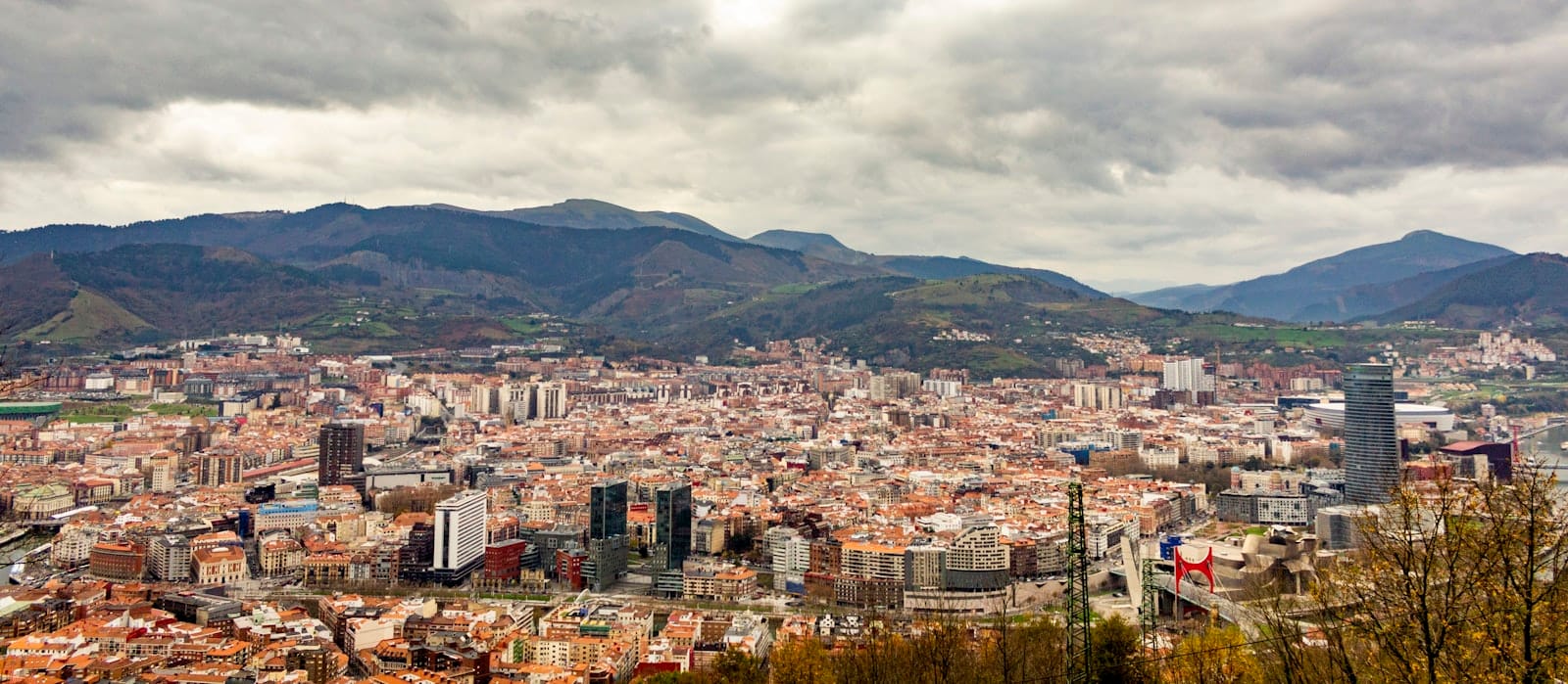 Panoramic view of Bilbao city under a dramatic cloudy sky with mountains in the background, conveying the mild winter atmosphere.