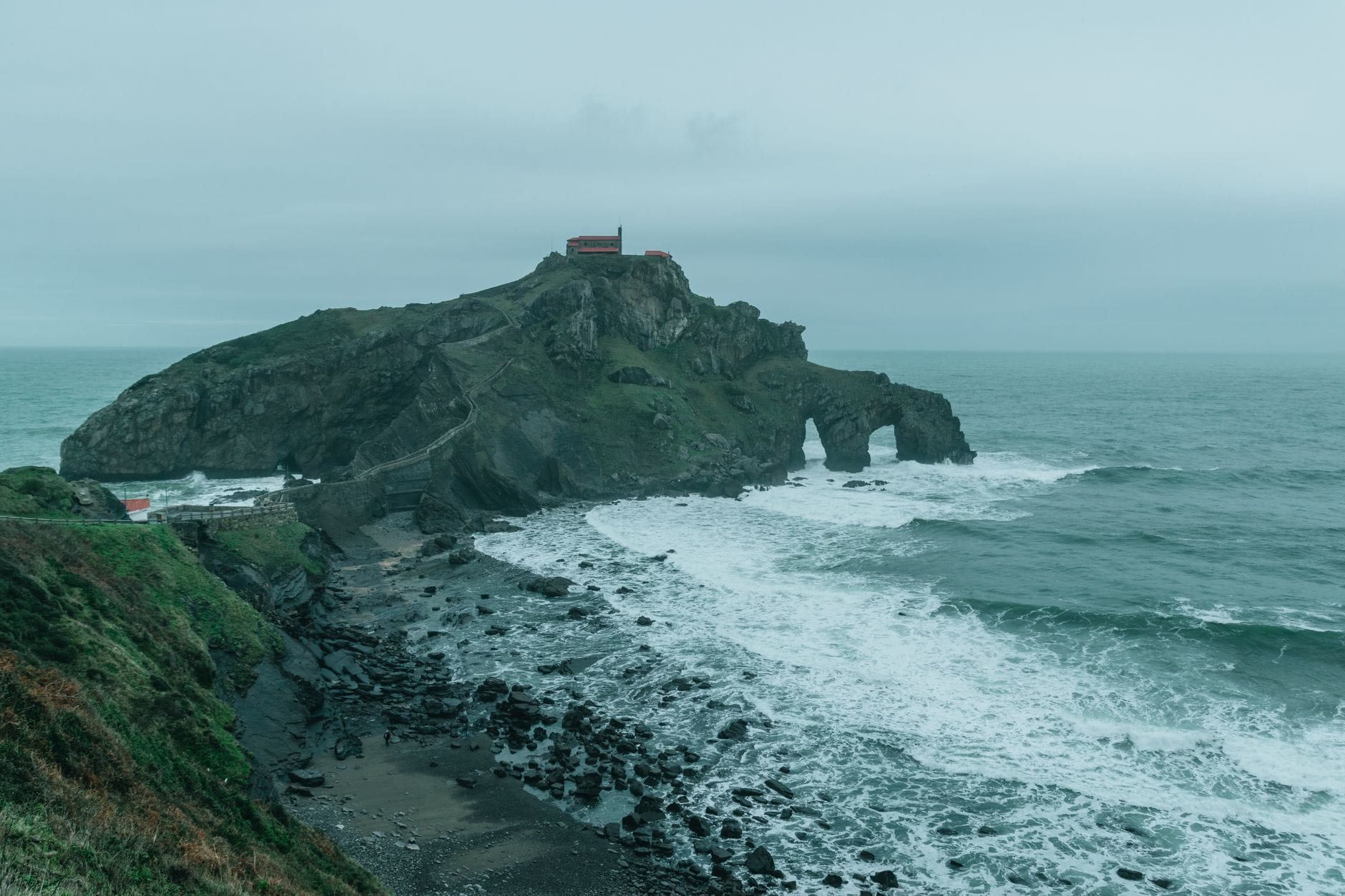 เกาะหินขรุขระพร้อมเส้นทางเดินเท้าที่คดเคี้ยวนำขึ้นไปยังโบสถ์น้อย คลื่นซัดเข้าหาชายฝั่งขรุขระใต้ท้องฟ้าครึ้มในฤดูหนาว